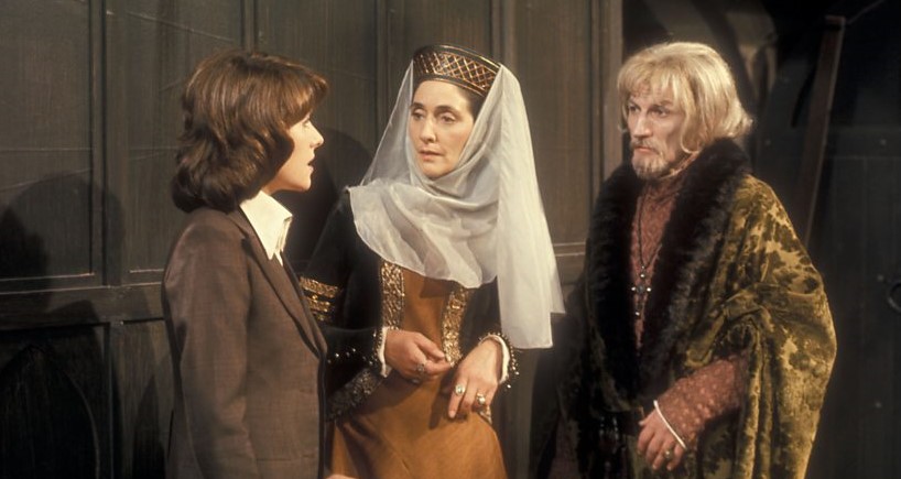 Left to right against a panelled wall and recess: Elisabeth Sladen as Sarah Jane Smith (a young woman in 1970s sports jacket and wing collared shirt), June Brown as Lady Eleanor (a mature medieval noblewoman in appropriate headgear and dress), and Alan Rowe as Edward of Wessex (a mature medieval nobleman in fur-lined gown, with white hair and beard, and a cross on a chain arond his neck).